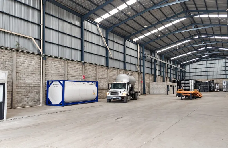 Camión blanco con tanque cilíndrico sobre la plataforma, dentro de bodega; a un lado hay otro recipiente en marco azul.