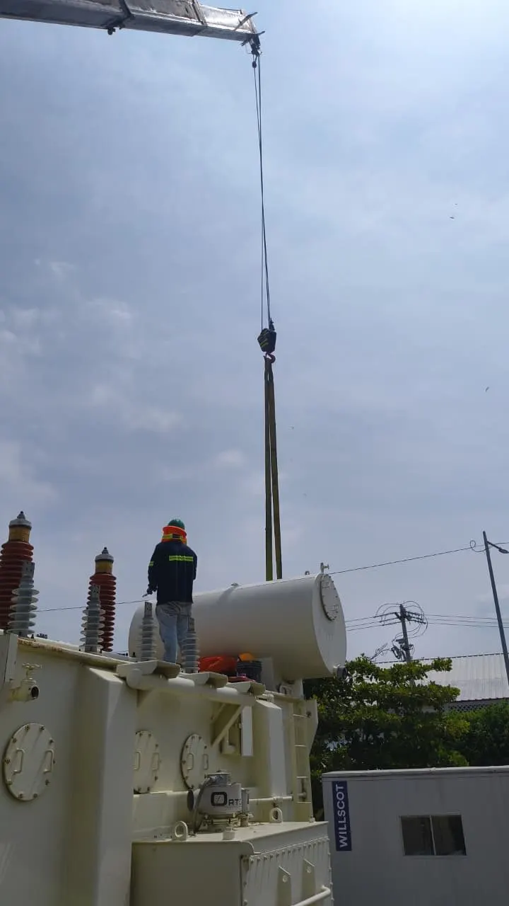 Camión grúa blanco junto a un transformador claro con radiadores y aisladores rojos en una instalación eléctrica.