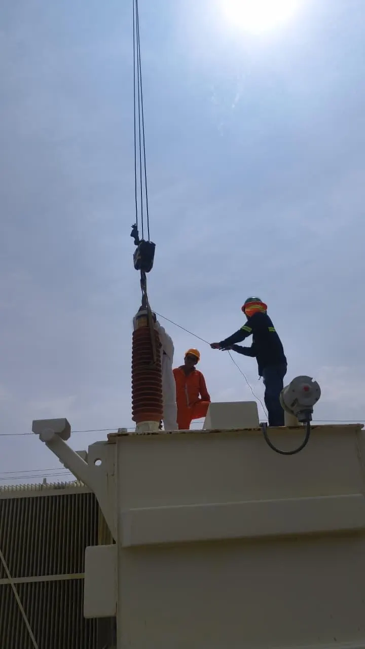 Tanque cilíndrico claro sujeto por eslingas sobre un transformador, con trabajadores en la parte superior.