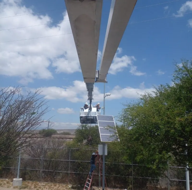 Vía férrea entre un camino y postes con paneles solares; cables aéreos y un trabajador con chaleco.