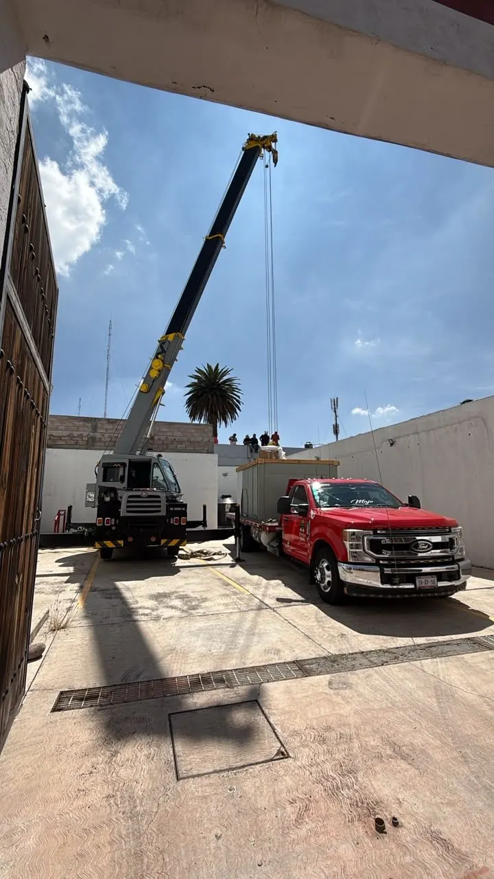 Grúa telescópica junto a un camión con paquete HVAC, dentro de un patio rodeado por muros.