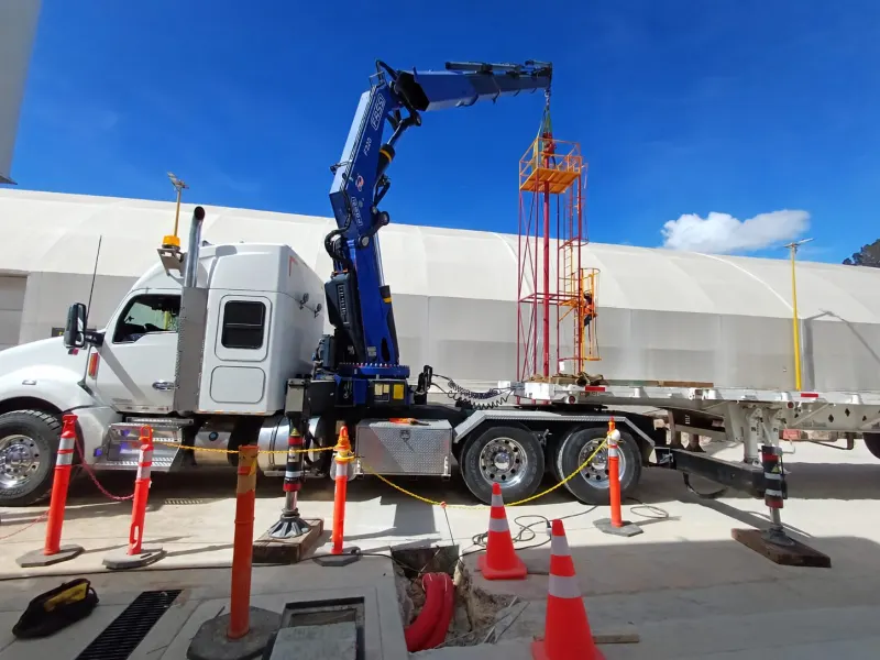 Camión con grúa articulada y estabilizador desplegado junto a una estructura metálica y tubería azul.