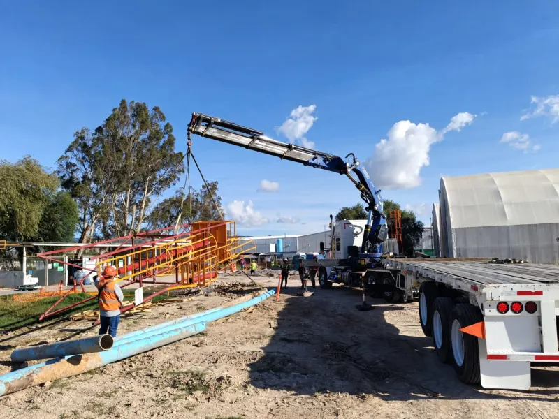 Grúa articulada azul sostiene una estructura metálica roja y amarilla junto a tubería azul y una plataforma.
